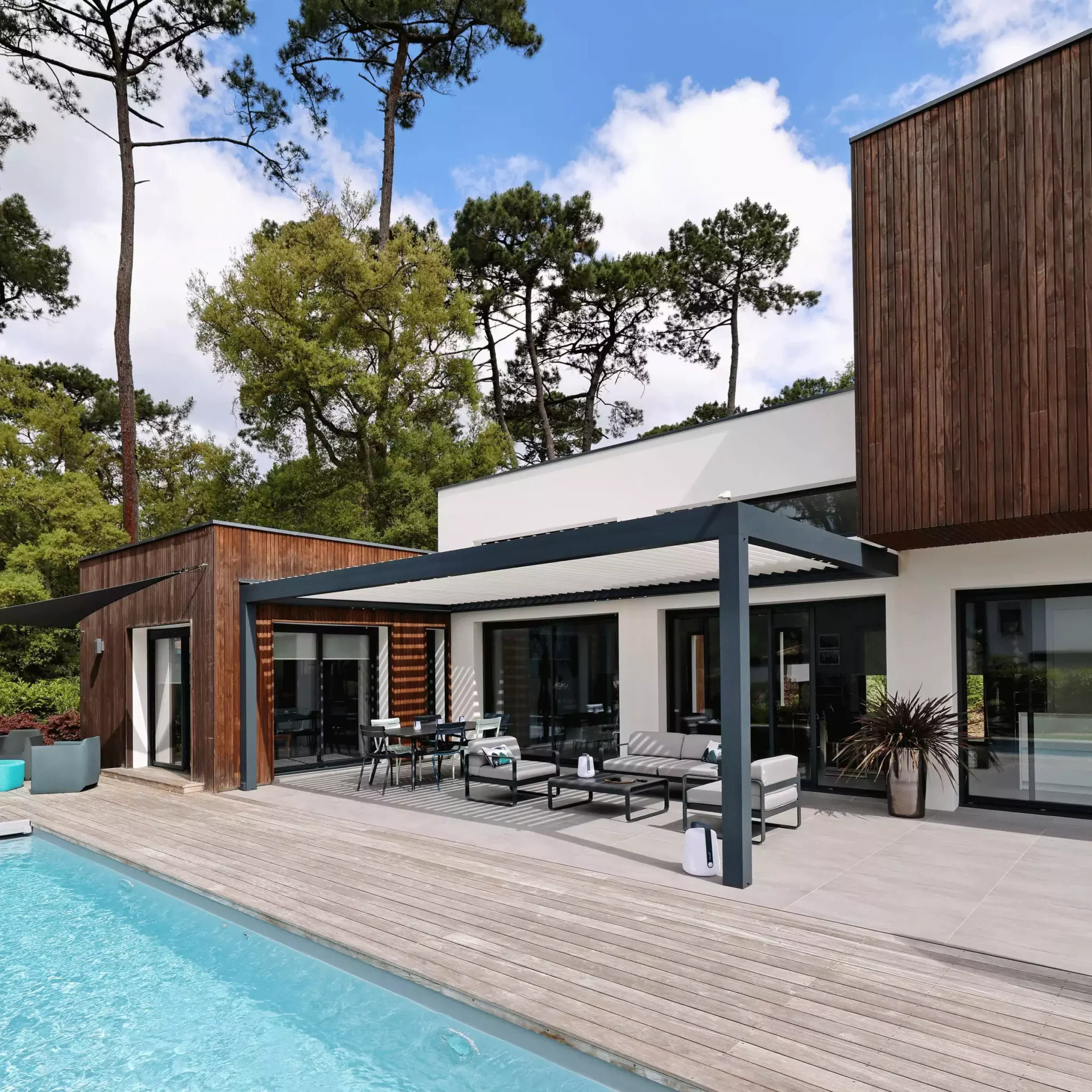pergola bioclimatique gris anthracite adossée à une maison d'architecte entourée de pins. Cette pergola abrite un salon de jardin moderne en métal.